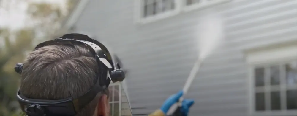 Technician in a face shield soft washing the siding of a house with a low-pressure wand